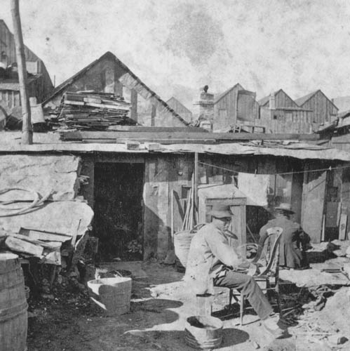 A Chinese resident of Virginia City's Chinatown seated outside a wooden dwelling among barrels and stacked lumber