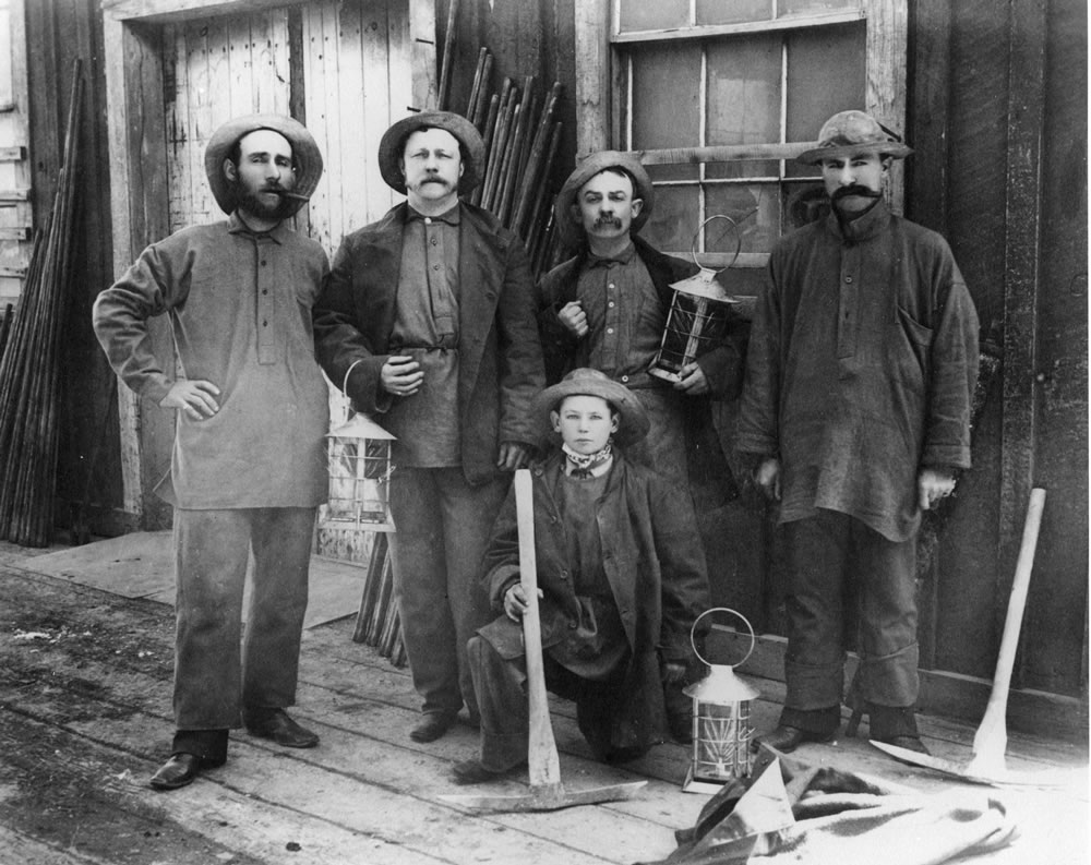 Four Comstock miners and a boy posing with lanterns and mining picks outside a mine building