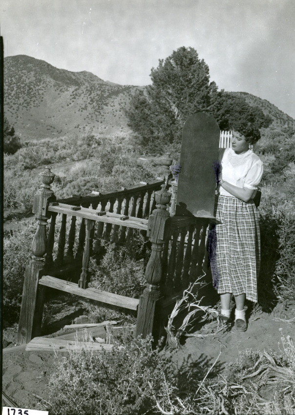 Woman standing beside a wooden grave enclosure on Flowery Hill, holding a carved J.B. headboard, circa 1949