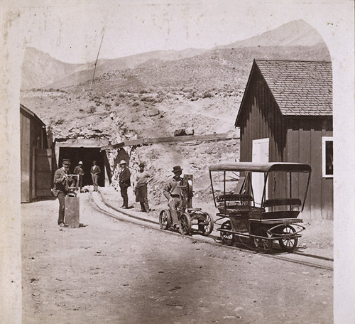Rail equipment and workers at the Sutro Tunnel portal complex, with mine buildings and hillside in the background