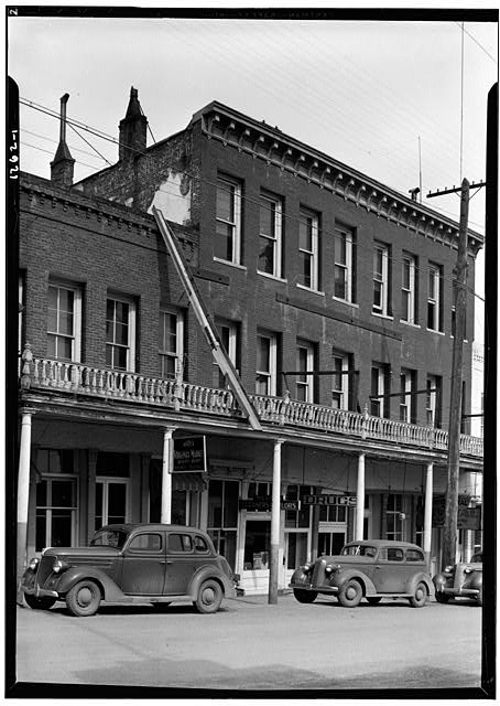Black and white 1937 street photograph of the three-story brick building housing Virginia City's Crystal Bar, with a columned sidewalk shelter and two parked cars