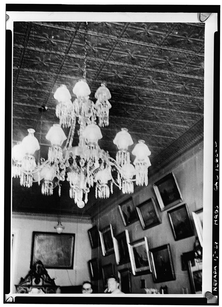 Black and white 1940 interior photograph of an ornate tiered crystal chandelier hanging from a pressed tin ceiling, with two men visible below